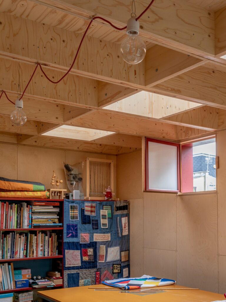 A cosy garden studio with wooden walls and ceiling, exposed beams, bookshelves, a patchwork quilt divider, hanging bulbs, and sunlight streaming through roof windows and an open red-framed window.