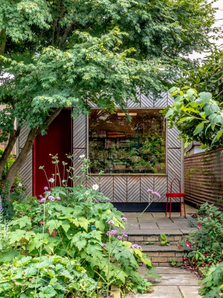 A small, modern garden studio with a large window is partially hidden by lush greenery and flowers. A single red chair sits by the entrance under the shade of a large tree, making this garden studio an inviting retreat nestled within nature.