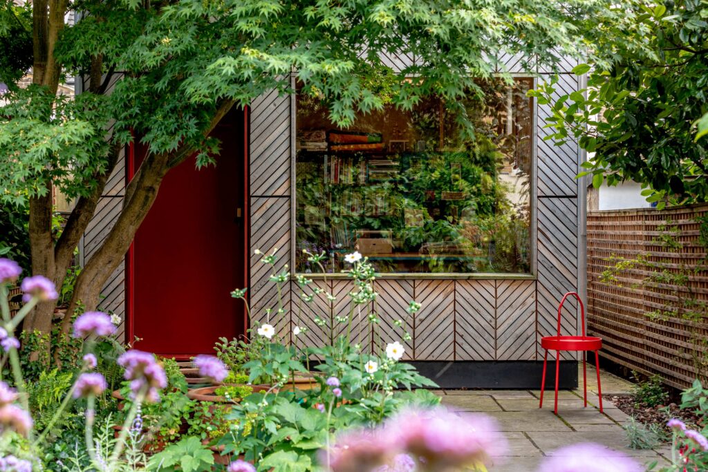 A small modern garden studio shed with a large window, red door, and red chair sits surrounded by green trees and blooming flowers in a lush back garden setting. Books are visible inside the window.