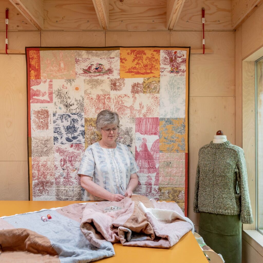 A woman works on fabric at a table in a craft room that feels like a cosy garden studio, with a colourful quilt on the wall behind her and a mannequin wearing a jumper and skirt by the window.