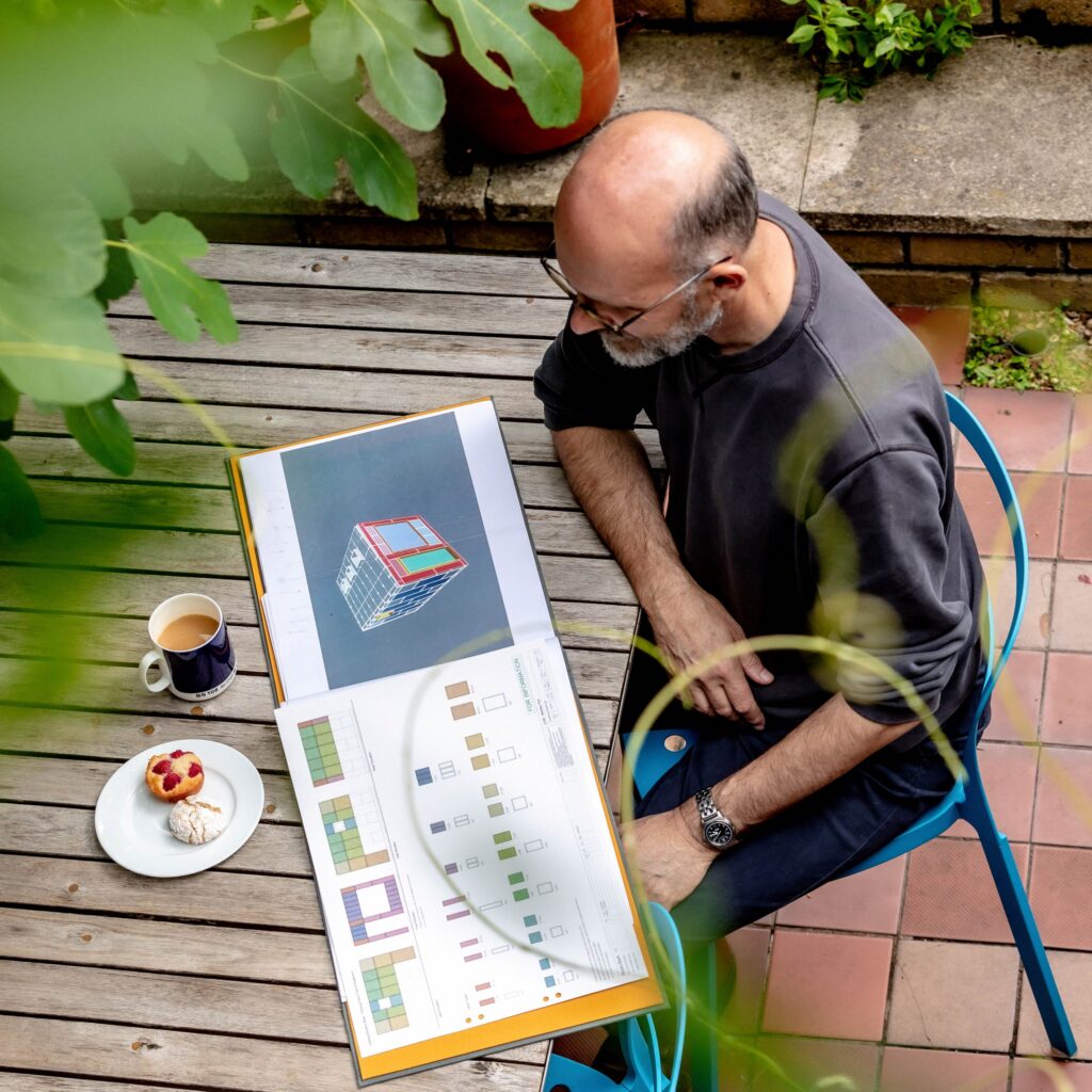 A man sits at an outdoor table in a garden studio, looking at architectural plans with diagrams and 3D renderings. On the table are a cup of coffee, a pastry, and a biscuit. Green plants and red tiles surround the scene.