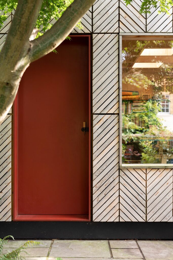 A red door and a window with a view of bookshelves are set in a wall with diagonal wooden panels, capturing the cosy charm of a garden studio. A tree branch partially frames the scene.