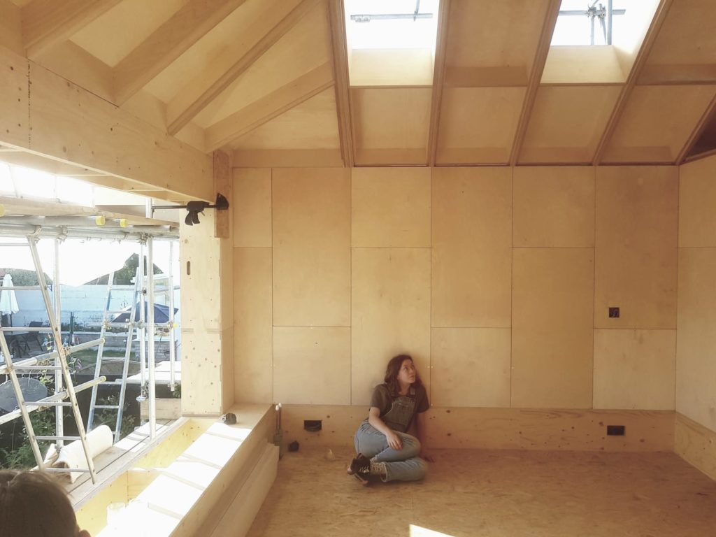 A person sits on the floor of a partially constructed timber room, leaning against the wall. Sunlight pours through large, open windows and unfinished beams above, highlighting the sustainable construction materials and scaffolding outside.