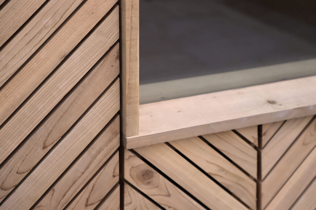 Close-up of a wooden garden studio structure with angled planks arranged in a chevron pattern. The studio features a rectangular opening framed by smooth, light-coloured wood, and the craftsmanship emphasises clean lines and geometric design.