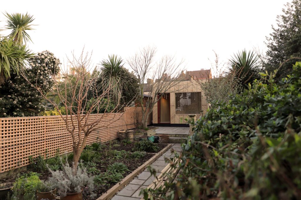A tidy back garden with a stone path, green plants, a wooden lattice fence, and a modern garden studio at the back. Trees and shrubs border the area, and the sky is clear.