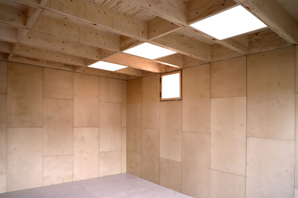 An empty garden studio with light plywood walls and ceiling, square skylights, and a small rectangular window. The floor also appears unfinished, matching the minimalist wooden interior.