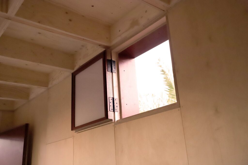 A small, square wooden window is open on a light wood-panelled wall, letting in daylight. The window, set within a garden studio, has a dark wooden frame and is hinged on one side.