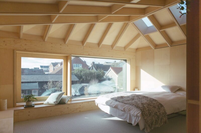 A bright, minimalist bedroom with wooden walls and ceiling, large windows overlooking rooftops, and a skylight. Sunlight streams in over a white bed. Cushions and a small plant sit on a window seat—ideal inspiration for bespoke projects.