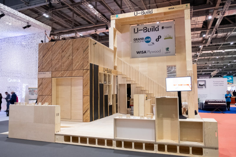 A modern, large wooden exhibition stand crafted from spruce with geometric patterns, signage reading U-Build, and display screens, set up in a spacious, well-lit event hall with other stands visible in the background.