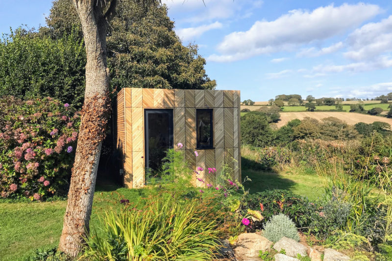 A small spruce garden shed with a chevron-patterned exterior sits among lush greenery, flowering plants, and trees, overlooking rolling fields under a partly cloudy sky.