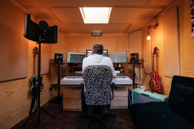A person in a white hoodie sits at a desk in a cosy music studio with birch guitars and recording equipment, surrounded by warm plywood and timber construction, facing multiple monitors and a keyboard.