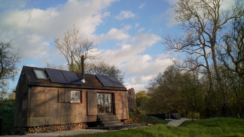 A small wooden cabin, reminiscent of a cosy Tiny House, with solar panels on the roof sits in a grassy area surrounded by leafless trees under a partly cloudy sky.