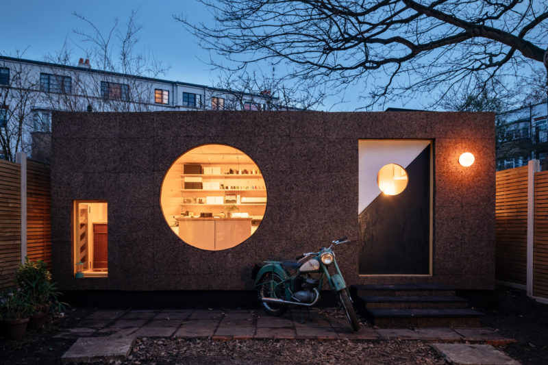 A modern, box-shaped tiny house with a large circular window, smaller square windows, and a geometric front door glows warmly at dusk. A vintage motorbike is parked outside, framed by leafless trees and sustainably sourced materials.