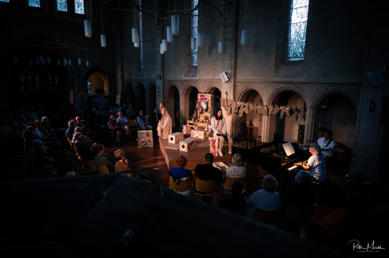 A stage performance takes place in a dimly lit church, where three actors stand near spruce-decorated modular boxes and a pianist plays. An audience watches as warm light streams in from the tall windows above.