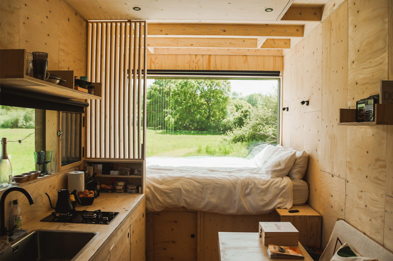 A cosy, modern tiny house interior with light wood walls made from sustainably sourced materials, a compact kitchen, and a bed next to a large window overlooking a green field. Natural light fills the space, creating a warm, inviting atmosphere.