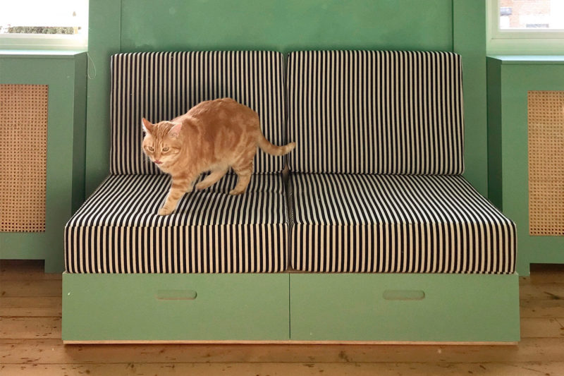An orange tabby cat walks across a black-and-white striped cushioned bench with green wooden sides and drawers, crafted from sustainably sourced materials, set against a matching green wall.