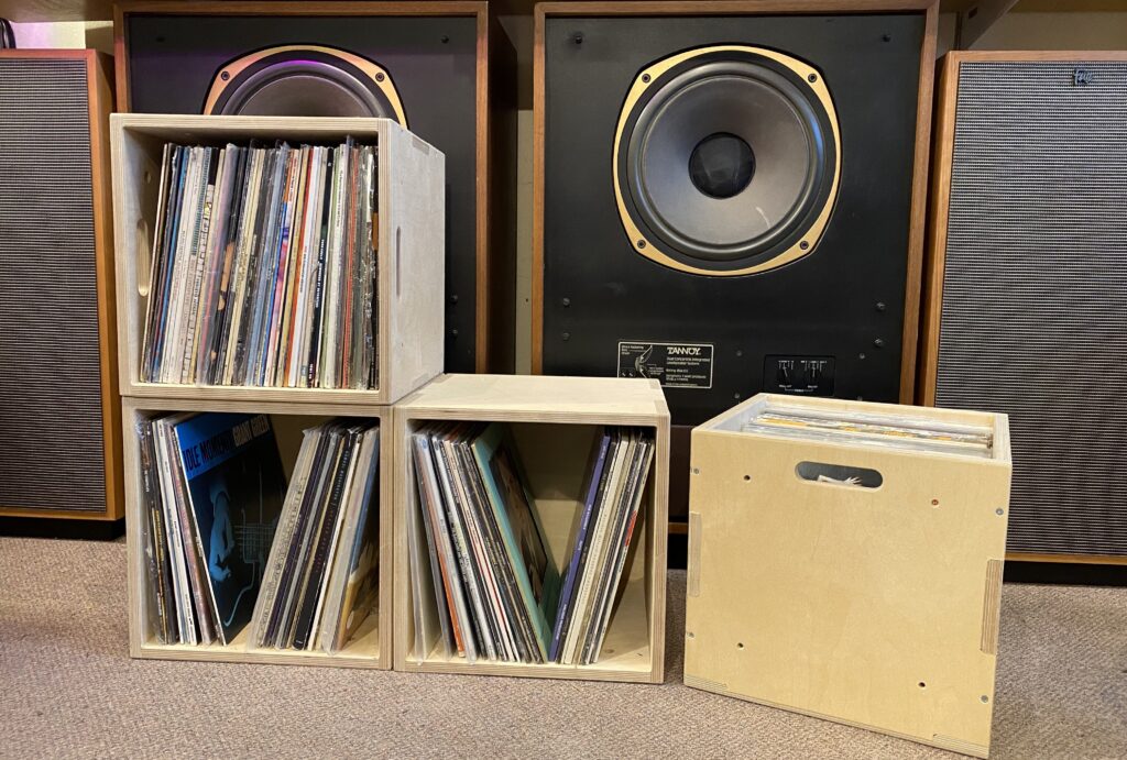 Four wooden U-Build cubes offer stylish vinyl storage, filled with records organised vertically in front of two large speakers. One crate features a handle cut-out. The scene, set on a carpeted floor, is perfect for any audio gold enthusiast.