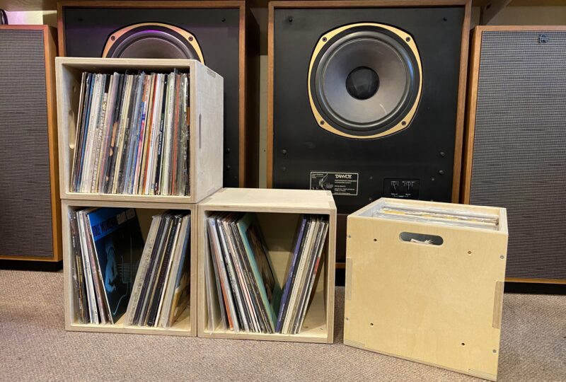 Four wooden U-Build cubes offer stylish vinyl storage, filled with records organised vertically in front of two large speakers. One crate features a handle cut-out. The scene, set on a carpeted floor, is perfect for any audio gold enthusiast.