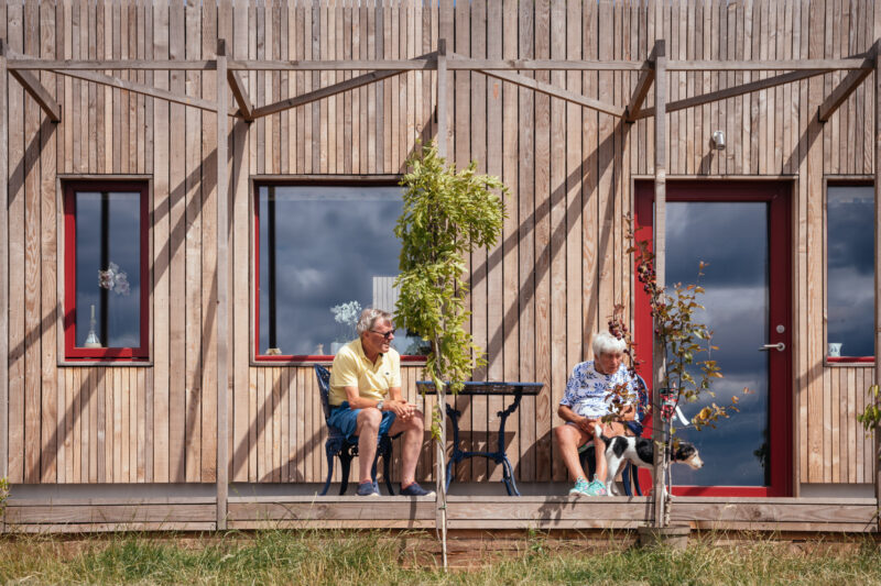 An elderly man and woman sit outside a wooden house, separated by a small table. The woman holds a dog on a lead. Both relax under the shade of spruce trees on a sunny day.