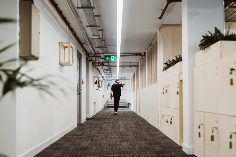 A person walks down a modern corridor with spruce lockers and plants on the right, white walls, modular design elements, and exposed ceiling pipes, heading towards a green exit sign at the end.