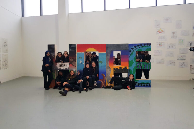 A group of students in school uniforms pose by large, colourful painted letters spelling “HOPE” in an art-filled classroom, celebrating sustainability through creative reuse of materials. Some students stand, while others sit or kneel on the floor.