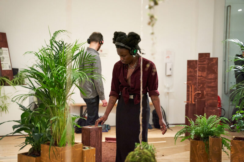 A woman wearing headphones stands among potted plants and modular wooden blocks indoors, appearing focused on her surroundings. A man in the background also wears headphones, both engaged in the immersive environment.