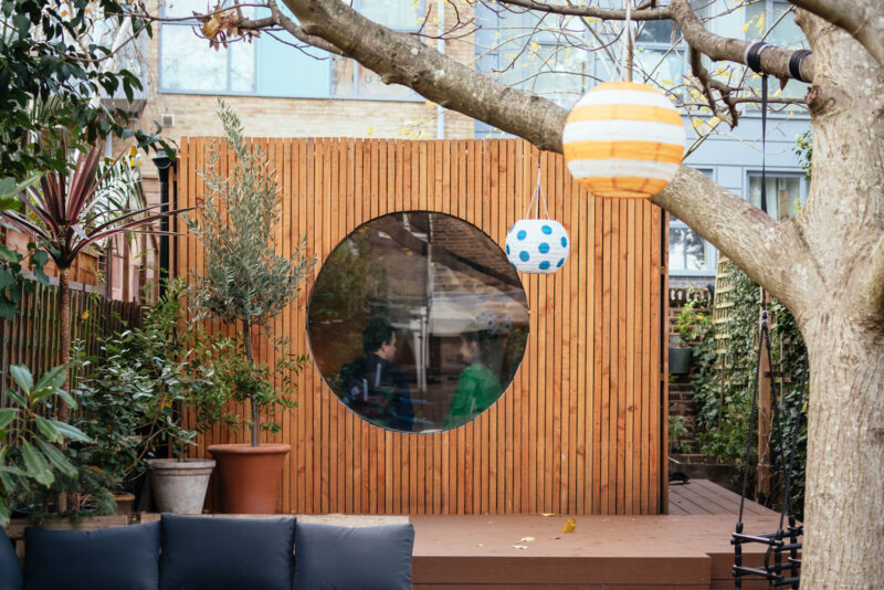 A cosy back garden features a spruce wooden wall with a large circular window, potted plants, and hanging paper lanterns in orange stripes and blue polka dots. Two people are visible through the window.