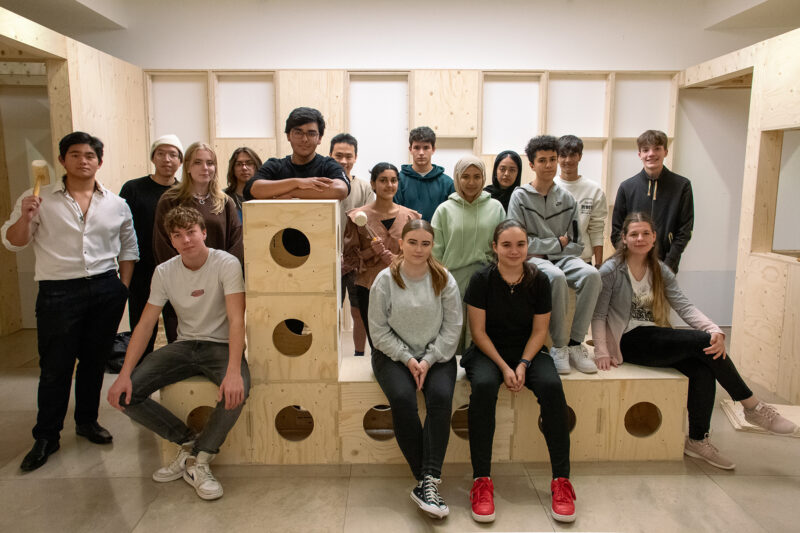A group of sixteen young people pose indoors on and around a large modular wooden structure with circular cut-outs, smiling and looking at the camera in a well-lit room crafted from sustainable spruce.