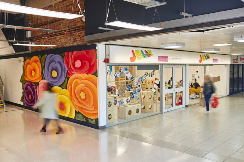A colourful shopfront with large, vibrant flower murals on the wall and glass doors labelled Jelly and making space for art. People walk by in a bright indoor corridor with polished floors and modular, sustainable design elements.
