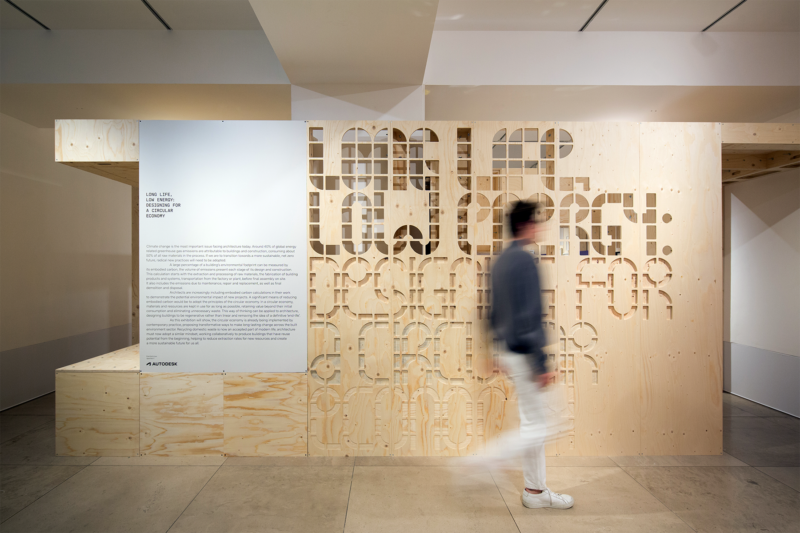 A person walks past a modular wooden wall with geometric cut-outs and a mounted sign containing text. The setting appears to be a modern gallery or exhibition space with clean lines and neutral colours.