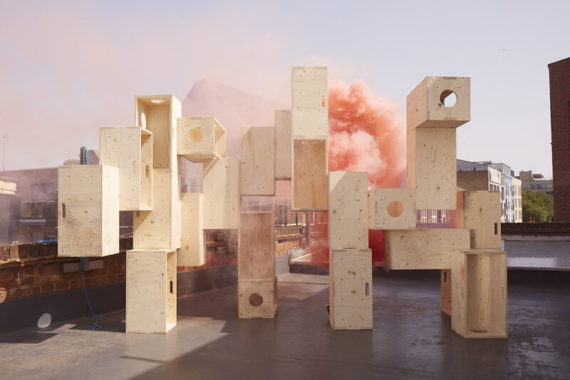 A group of large, light plywood boxes is stacked in an abstract arrangement on a rooftop, with orange smoke billowing behind them and buildings visible in the background under a clear sky.