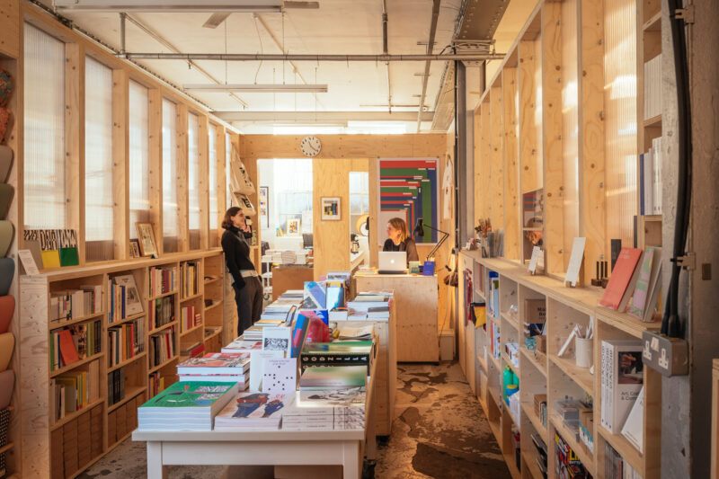 A cosy, well-lit bookshop with sustainable birch wooden shelves full of books and magazines, tables displaying colourful books, and two people talking at the counter in the background under a wall clock.