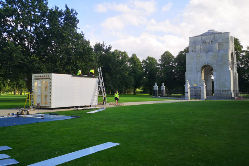 Workers are assembling a white modular structure using ladders in a park with green grass. Nearby stands a large stone arch monument, surrounded by trees under a partly cloudy sky.