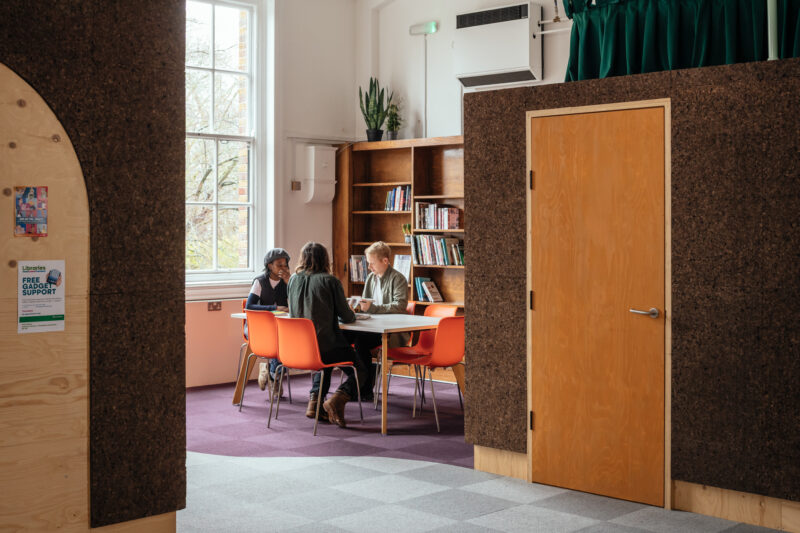 Three people sit around a table in a library or study room with birch bookshelves, orange chairs, and natural light streaming in. A wooden door and a poster are visible in the modular space’s bright foreground.
