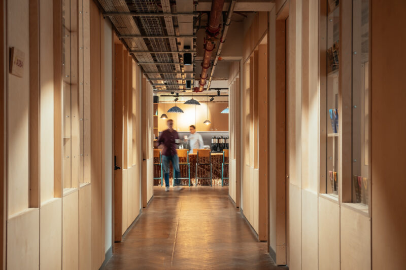 A modern office corridor with birch plywood walls and ceiling pipes leads to a well-lit communal area. Two people are visible in the background, one walking and one standing near bookshelves and hanging lights.