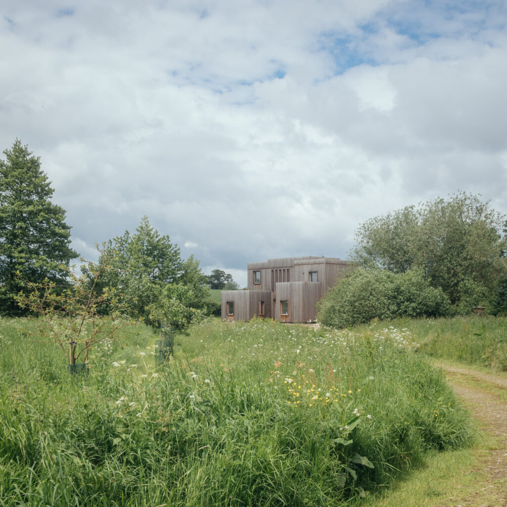 A modern wooden house built with the U-Build system sits among tall grass and wildflowers, surrounded by trees under a partly cloudy sky. A dirt path curves through the greenery towards the building.