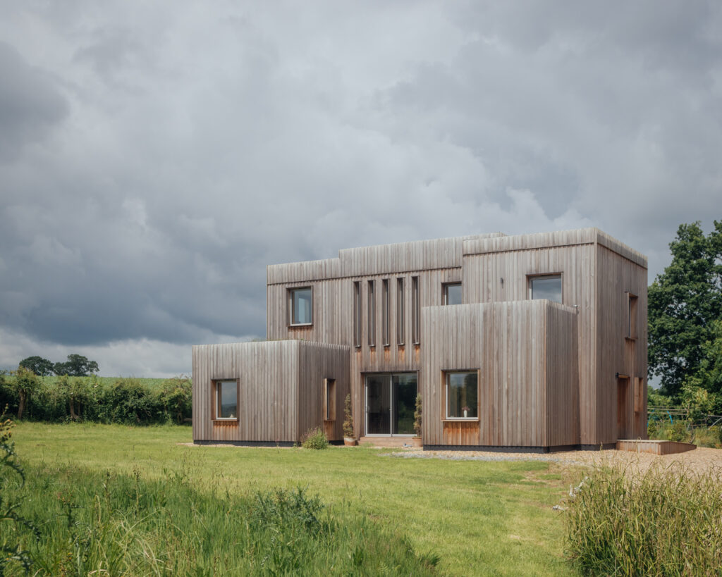 A modern two-storey house with a wooden exterior, large windows, and a flat roof stands in a grassy field under a cloudy sky. Featuring plywood construction, shrubs and trees are visible in the background.