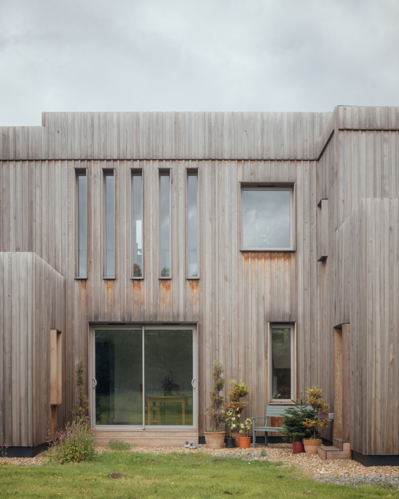 A modern two-storey house with vertical wooden slats, large windows, and a sliding glass door showcases sleek plywood construction. Potted plants and small trees frame the entrance, with grass and gravel in the foreground under a cloudy sky.