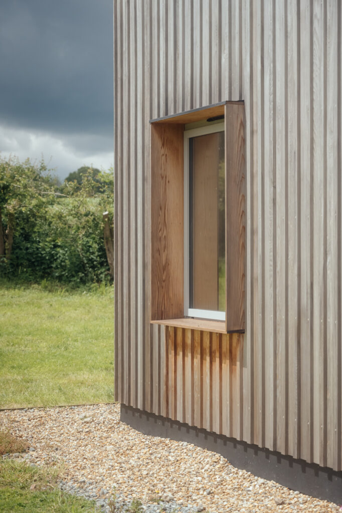 A modern wooden house built with vertical slats and a single rectangular window, surrounded by grass and gravel, with cloudy skies and greenery in the background—showcasing sleek plywood construction.