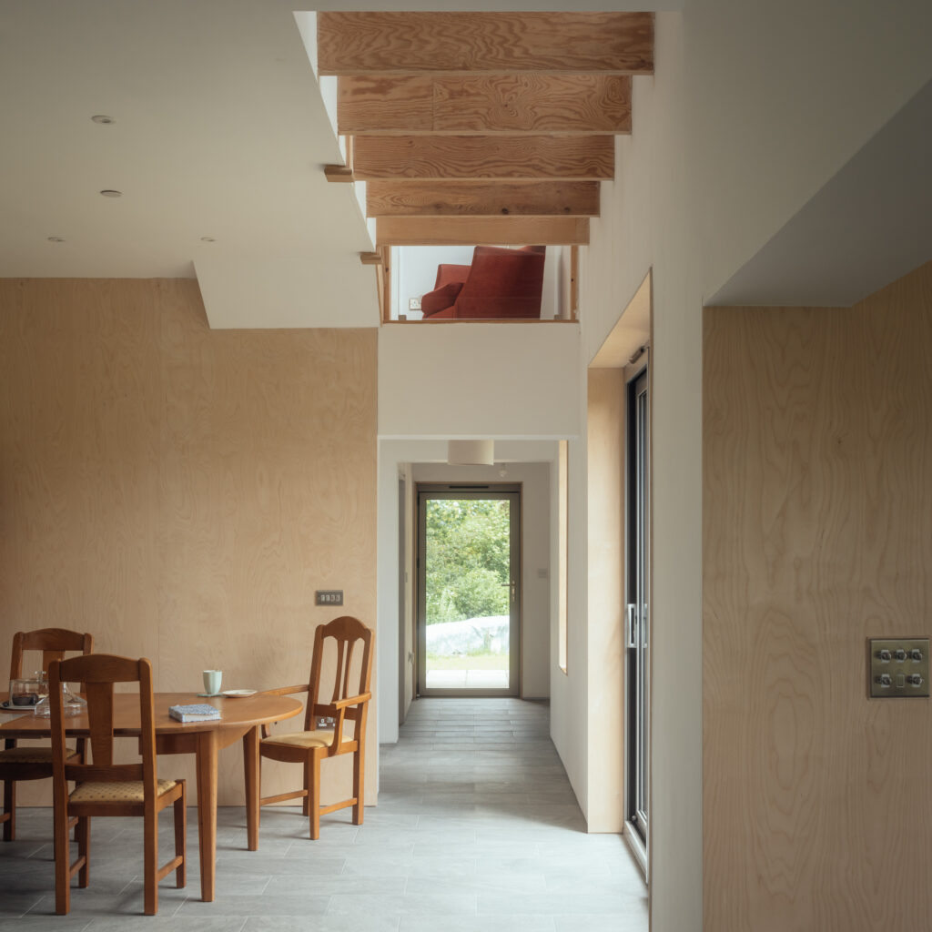 A modern interior with wooden furniture, a round dining table, chairs, light wood walls in plywood construction, a hallway with a glass door at the end, and a loft space above featuring a red armchair. Natural light fills the house.