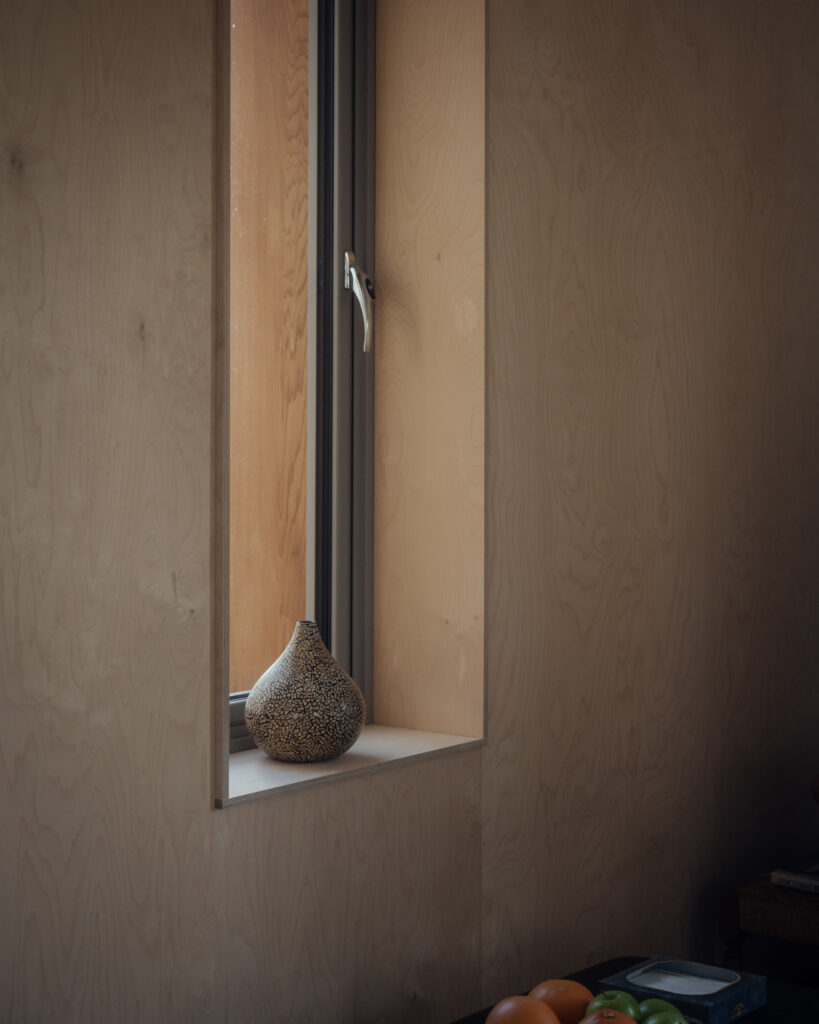 A patterned ceramic vase sits on a white windowsill in a minimalist room with light wood-panelled walls, highlighting plywood construction. A partially open window lets in soft daylight. Some fruit rests on a dark surface in the lower right corner.