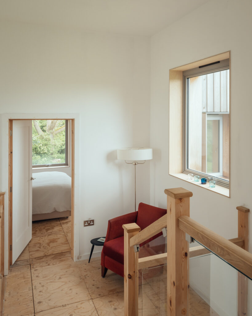 A bright hallway with wooden floors and stair railing built using the U-Build system, featuring a red armchair, small round black table, white floor lamp, and large window overlooking greenery. An open door leads to a bedroom.