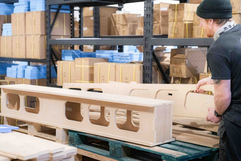 A person assembling or inspecting a large modular wooden box with several rectangular cut-outs in a warehouse filled with stacked cardboard boxes and shelves.
