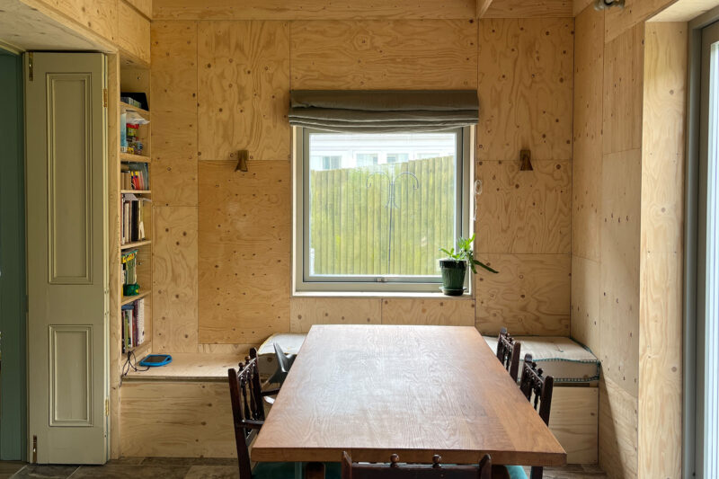 A cosy dining nook with birch wooden walls, a wooden table and chairs, bench seating under a window with a potted plant, wall lamps, and a built-in bookcase to the left. Natural light fills the sustainable space.