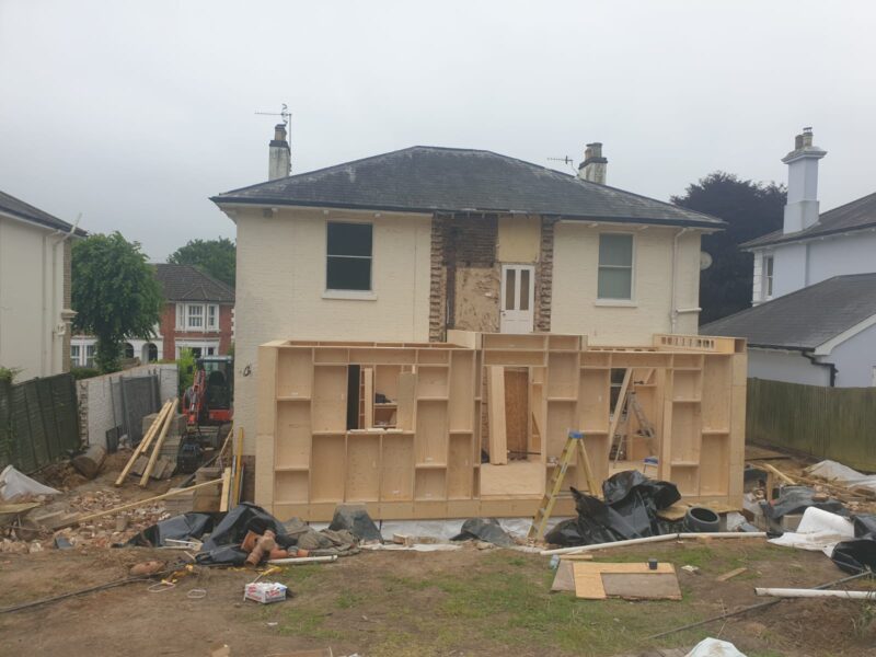 A two-storey house with construction work in progress, featuring a sustainable timber construction framework being built at the back. Building materials, tools, and debris are scattered around the building site.