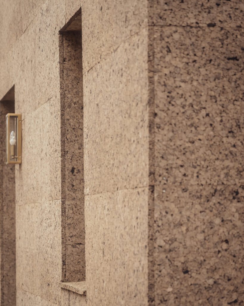 A wall covered in textured, tan cork cladding with a vertical recessed niche and a gold-toned wall light.