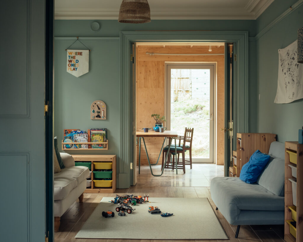 A playroom with green walls featuring a wall hanging that says Where the wild ones play, a low bookcase, and a rug with toy vehicles, with timber double doors looking out to a dining area and patio.