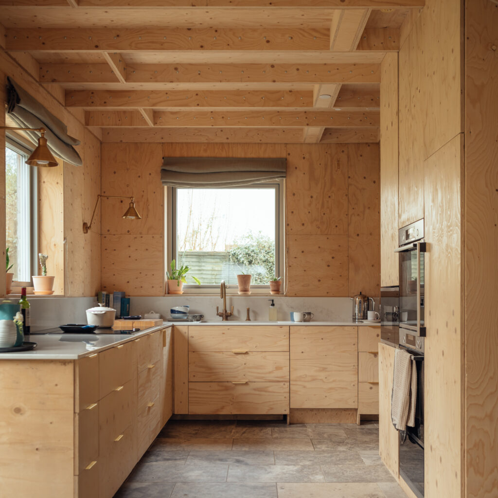 Modern kitchen with light plywood cabinetry and exposed timber beams, featuring a sink, worktops, and a wall-mounted oven.