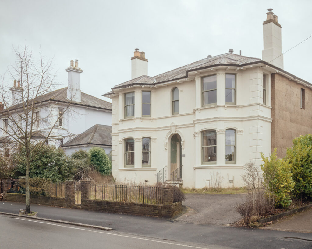 A two-storey, cream-coloured house with a bay window and timber cladding, set behind a brick wall and metal fence, with a similar house visible next door.