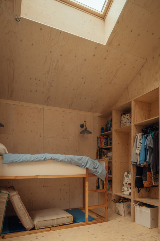 A bunk bed in a plywood-clad, timber room with a skylight, featuring a light blue blanket, open shelving, and a hanging clothes rail.